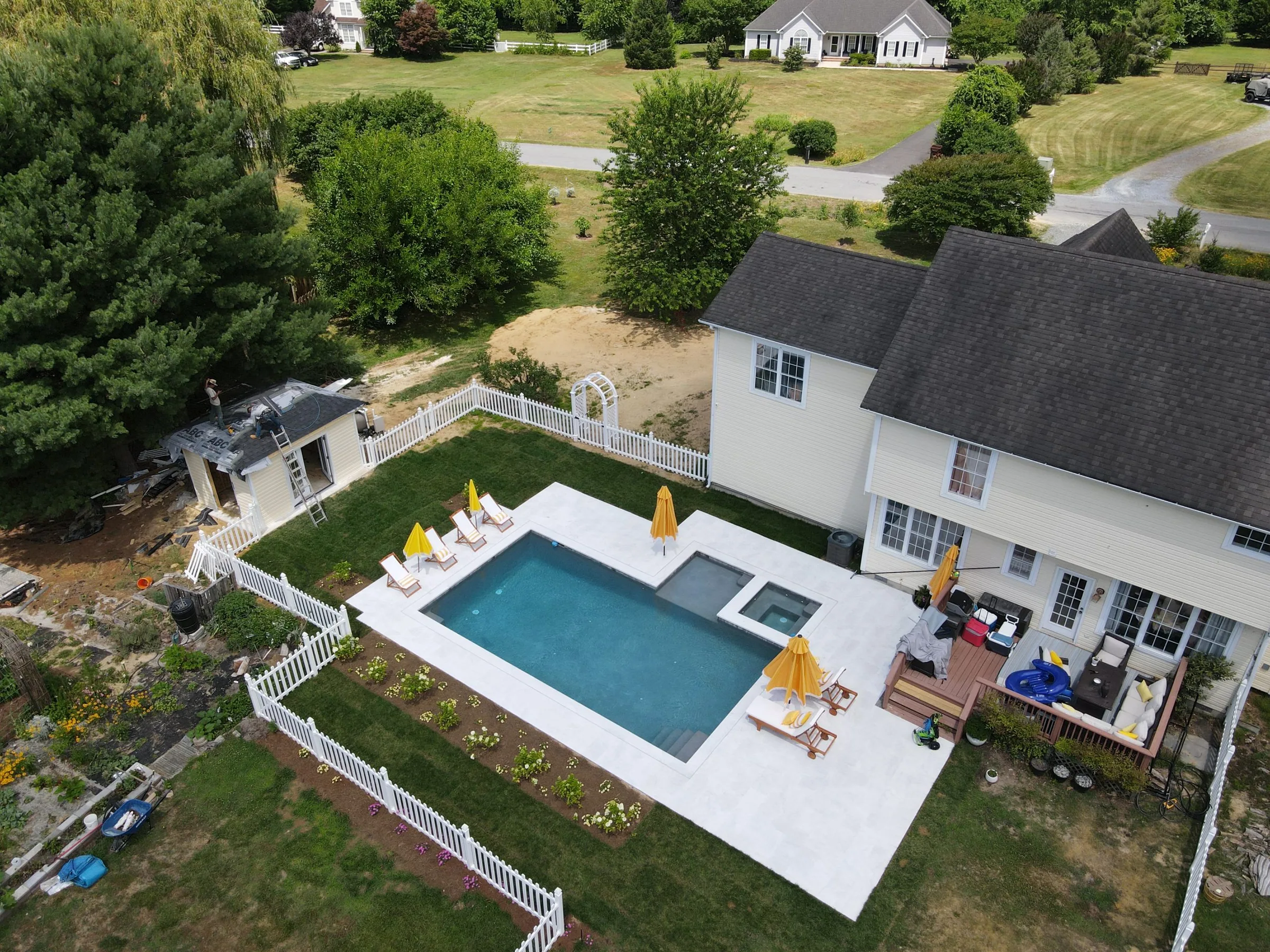 Aerial view of a backyard pool, white stone patio, and surrounding lawn
