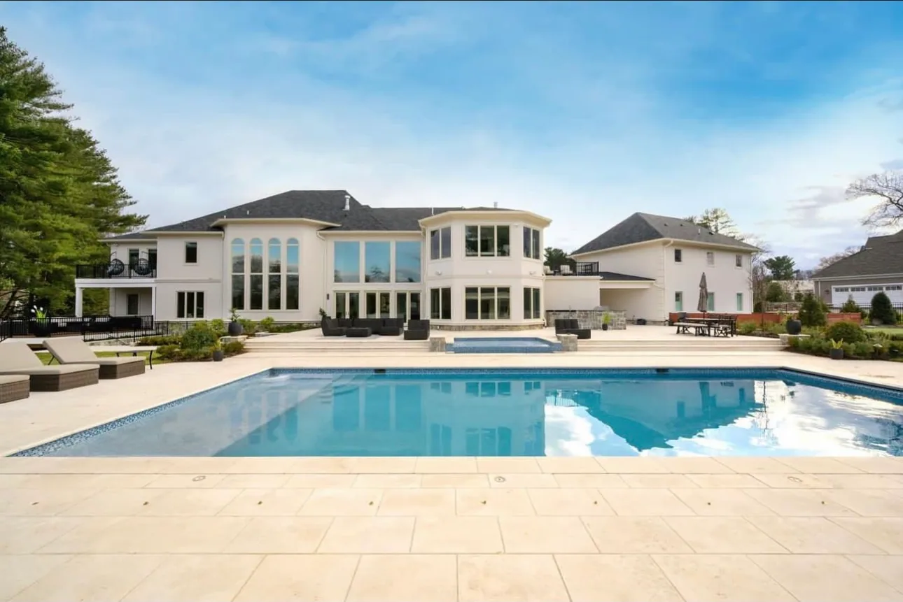 Swimming pool set into a large paved terrace beside a white house
