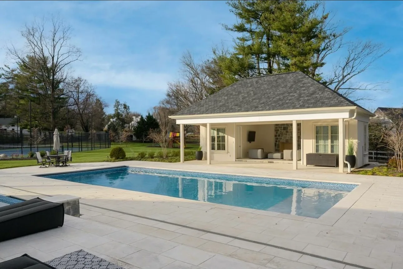Rectangular pool and stone patio with a pool house and lawn beyond